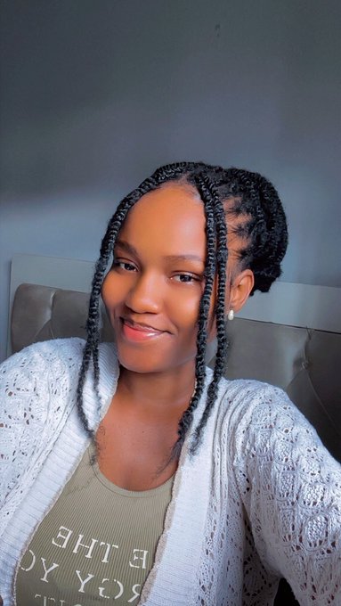 A selfie of a smiling young Black woman with long braided hair wearing a light grey knit cardigan over an olive shirt with partially visible text 'E THE' and 'US OY'.