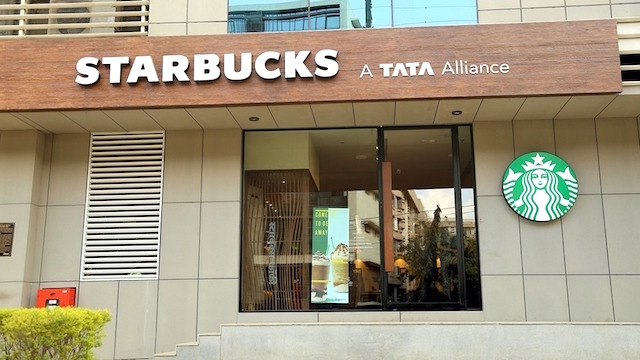The image shows the exterior of a Starbucks café featuring signage that reads "STARBUCKS A TATA Alliance," with the iconic green Starbucks mermaid logo mounted on the right side of the entrance. The Tata Alliance branding highlights Starbucks' joint venture with the Indian conglomerate Tata, contextualizing the news of Starbucks expanding its operations by establishing a Global Capability Centre (GCC) in Chennai, India.