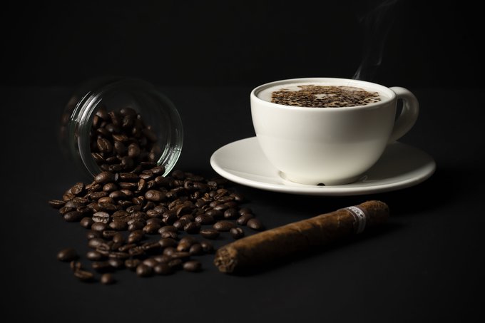 A still life composition features a white cup of steaming coffee with a rich crema, positioned next to a tipped glass jar spilling roasted coffee beans and a brown cigar on a dark surface.