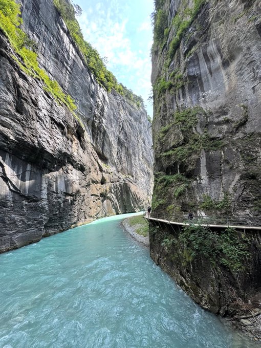 A scenic view of a turquoise water river flowing between tall, sheer rock canyon walls with a narrow walkway carved into the cliffside where tiny figures of people can be seen.