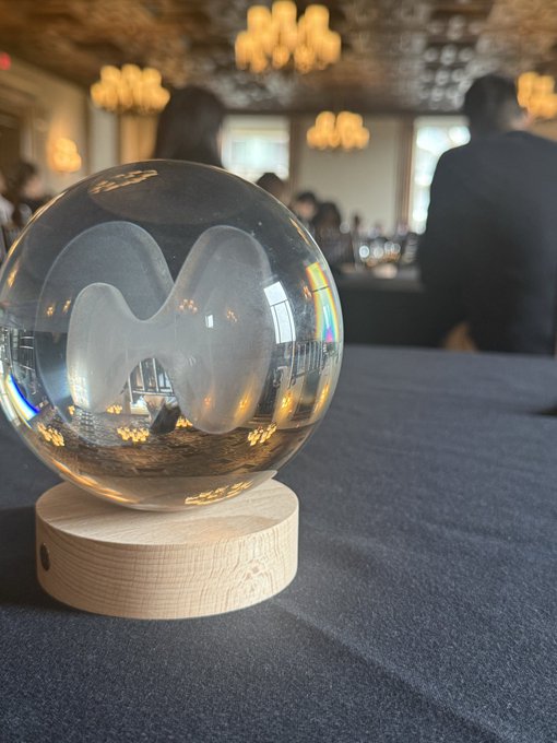 A clear crystal ball with an etched 'M' rests on a wooden base on a dark tablecloth in an elegantly lit room with chandeliers.