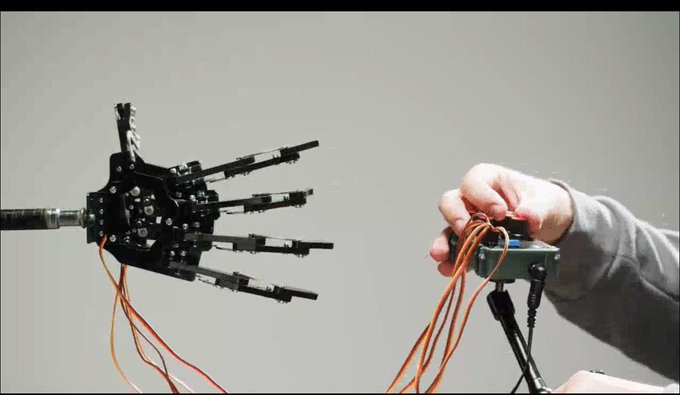A person wearing a gray long-sleeve shirt holds a black device with orange wires connected to a robotic hand, demonstrating how human actions can be captured to train Physical AI systems. The robotic hand's articulated black fingers mirror the human grip, illustrating how natural movements become valuable training data for AI development.