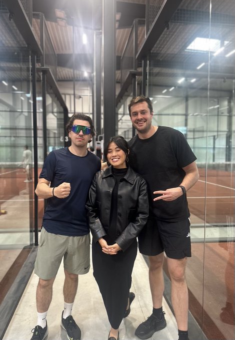 The image features three people standing together, posing for a photo. They are all dressed casually, with one person wearing a black jacket over a white shirt. The background of the picture shows an indoor setting with glass walls that allow natural light to enter the room. A tennis court can be seen in the background as well. The caption of the image reads: "guten morgen from berlin on my way back from the Berlin Solana Summit! might've been the biggest solana event in europe so far! Our @craftsdev also had a lightening talk on  why tokens are broken and how to fix them".