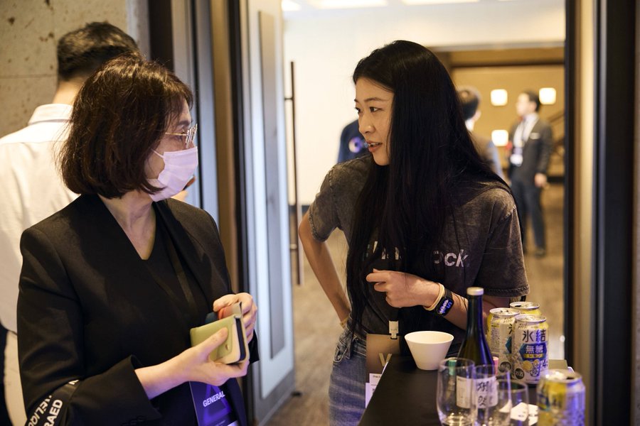 Two women converse at an indoor event; one wears a mask and holds a phone while the other wears a grey 'Hard Rock' t-shirt near drinks including a can with Japanese text.