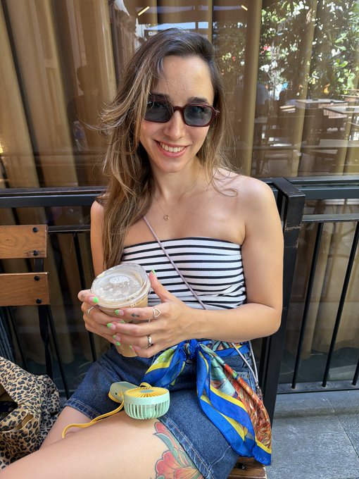 A smiling woman with sunglasses and a black-and-white striped top sits outdoors holding an iced coffee, with a portable fan in her lap.