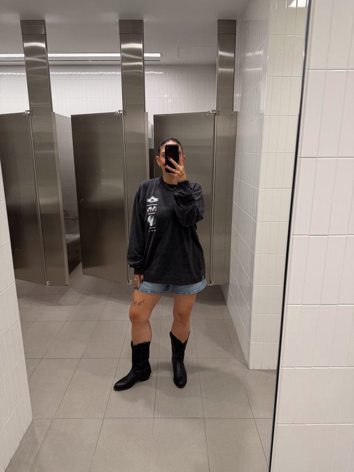 A mirror selfie of a person in a public restroom stall, wearing an oversized dark blue long-sleeve shirt, denim shorts, and black mid-calf boots.