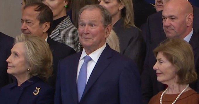 A photograph of George W. Bush standing in a crowd at a formal event, flanked by Hillary Clinton on the left and another woman on the right.