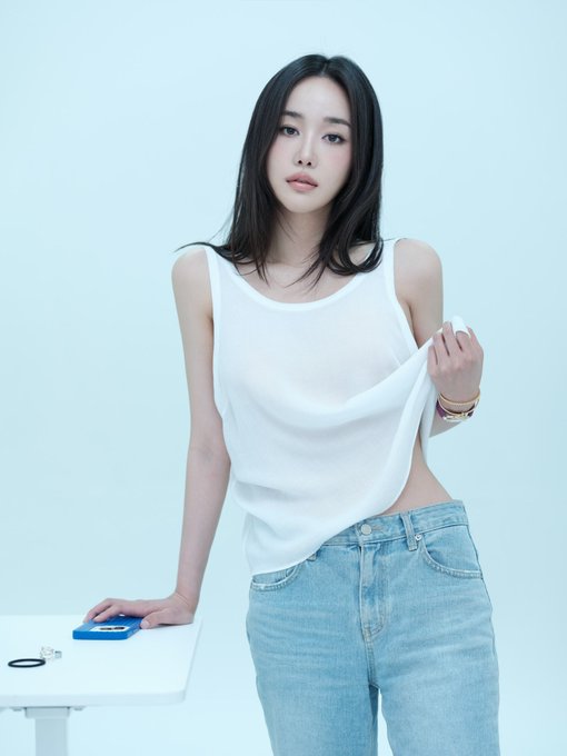A portrait of a young woman with dark hair wearing a white sleeveless top and light blue jeans, posing against a light blue background with one hand resting on a white table holding a small blue object.
