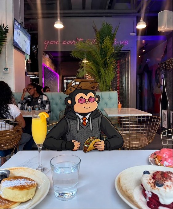 An image of a cartoon monkey character wearing sunglasses, a black hoodie, and a Hogwarts-style hat, sitting at a restaurant table with pancakes, a mimosa, and other brunch items, edited onto a real restaurant photo.