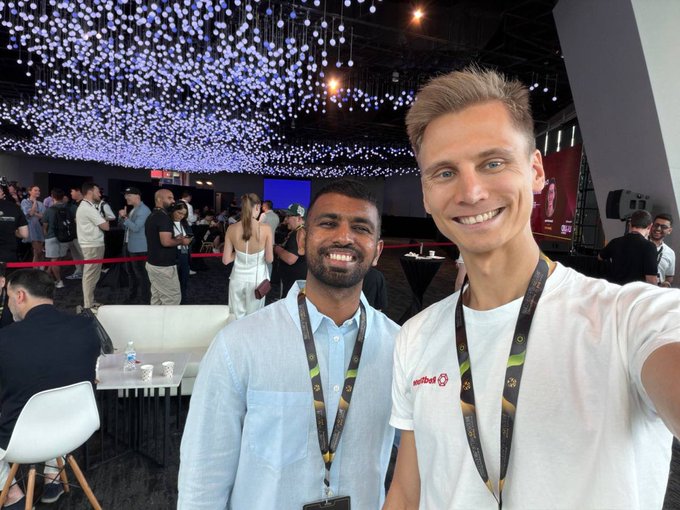 two men taking a selfie at a conference event under a ceiling of hanging decorative lights, both wearing lanyards with badges