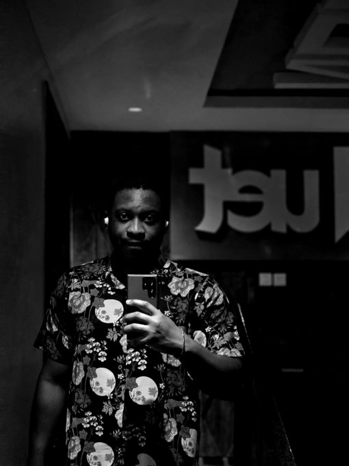 A black and white selfie shows a man wearing a dark shirt patterned with sugar skulls, holding a smartphone, with partially visible stylized text reading "au" on the wall behind him.