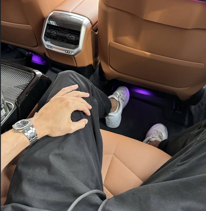 The image shows a person sitting inside a car, with their feet resting on the floor of the vehicle. They are wearing black pants and white shoes. The car has leather seats that match the color of the person's shoes. In front of them is an air freshener, which appears to be purple in color.