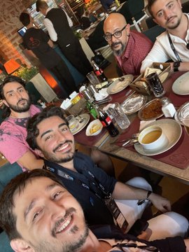 The image shows a group of five men sitting around a dining table at a restaurant. They are all smiling for the camera while enjoying their meal, which includes various dishes such as soup, bread, and other food items. The atmosphere appears to be casual and friendly, with everyone engaged in conversation and having a good time together.