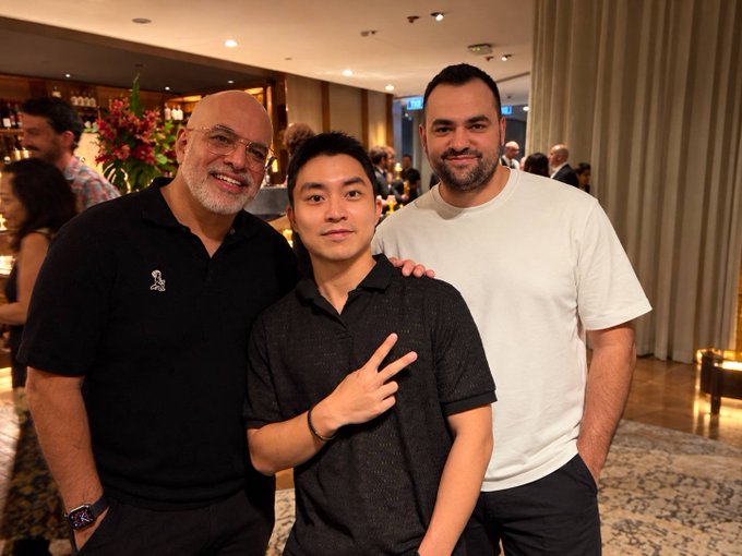Three men pose together at an indoor event, with the man on the left wearing a black polo with a logo, the center man making a peace sign in a dark polo, and the right man in a plain white t-shirt.