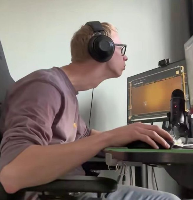 A side-profile shot of a young man wearing glasses and black headphones, focused on a computer monitor displaying an orange-toned image, with a microphone visible nearby.