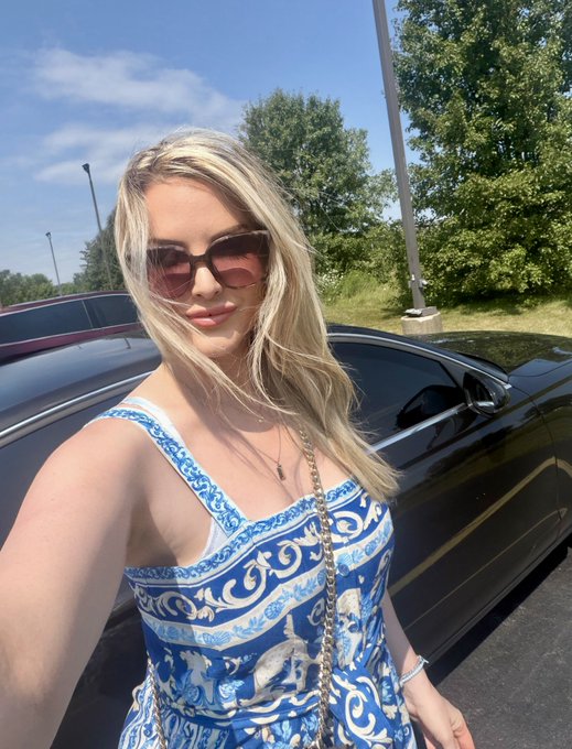 A blonde woman wearing sunglasses and a blue and white patterned dress poses in front of a black car outdoors.