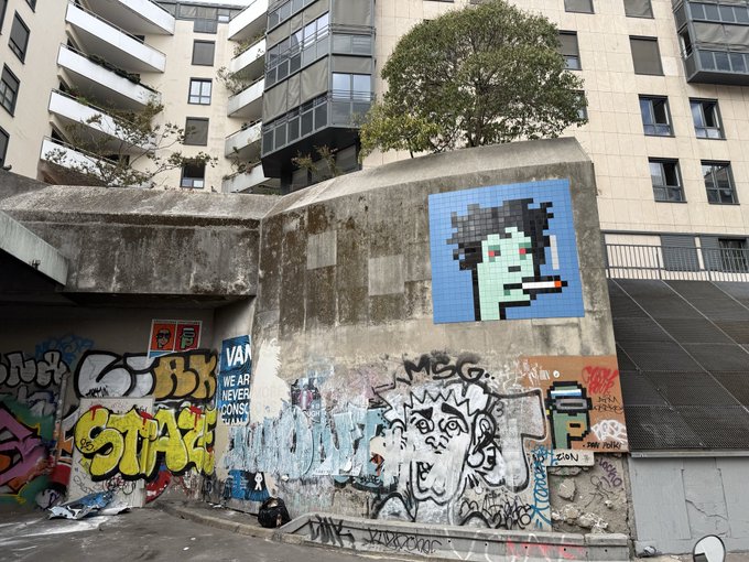 An urban alley scene features a concrete wall covered in graffiti, including a pixel-art mosaic of a green-faced character, set against apartment buildings and a tree.