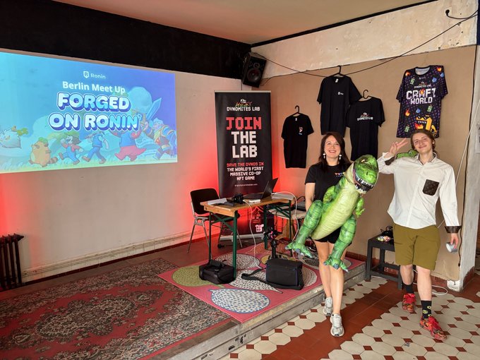 The image shows two women standing next to a table with a large green dinosaur balloon, possibly a LEGO dinosaur or a stuffed animal. They are positioned near a screen displaying "Berlin Meet Up Forged On Ronin" along with an advertisement for a gaming event. The room has a tiled floor and a projector screen mounted on the wall behind them. There is also a banner hanging from the ceiling in the background, which reads "work in progress see you later at @Ronin_Network Crypto Gaming Meet Up".
