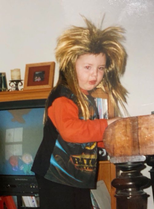A young child poses indoors wearing a voluminous, wild blonde wig and an orange shirt under a black vest, standing near a wooden staircase post and a television.