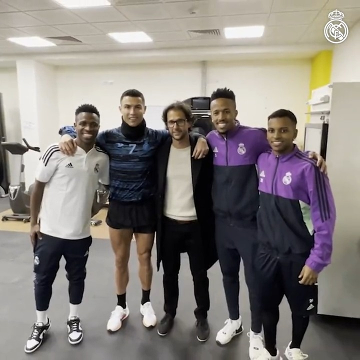 Cristiano Ronaldo poses with three other men, including a man in a dark coat, in what appears to be a gym setting with a Real Madrid logo in the corner.