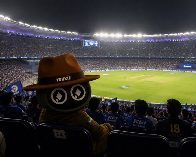 The image shows a large stadium filled with people watching a cricket match at night. In the foreground, there's an unusual mascot wearing a hat that says "Youkie" along with two eyes. The mascot appears to be sitting on a chair in front of the crowd, and it seems like they are enjoying the game together. The image also contains text about DeFi and liquidity issues, which may not have any direct connection to the cricket match or the mascot's presence at the stadium.