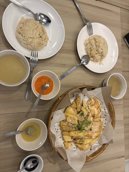 An overhead view of a meal featuring plates of rice, a basket of sliced chicken, and dipping sauces including a bright red chili sauce and a clear broth.