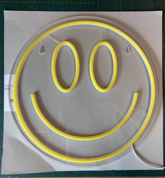 A yellow neon smiley face sign with oval eyes is mounted on a clear circular acrylic backing, sitting on a green cutting mat.