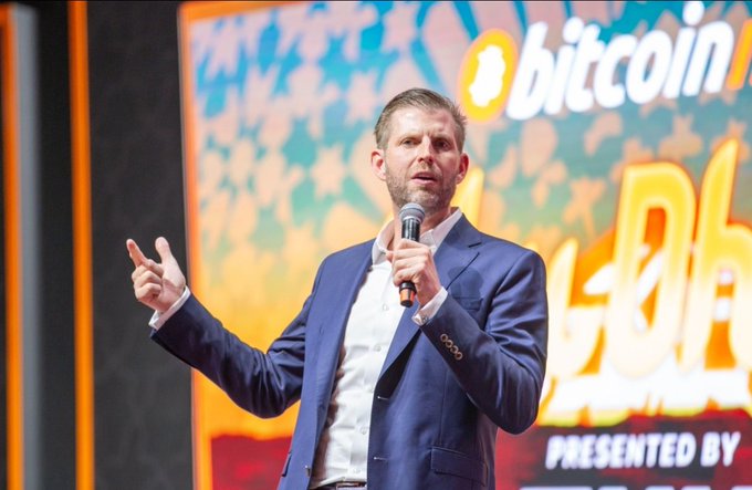 Eric Trump speaks into a microphone at the Bitcoin 2024 conference, where his "$1 million BTC" prediction echoes on stage. The backdrop displays the event's "bitcoin" logo, tying his bullish outlook to the broader crypto narrative highlighted in the post.