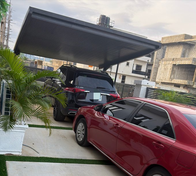 A black SUV and a red sedan are parked side by side under a metal carport in a residential driveway, with a palm plant and surrounding buildings visible. The image likely represents the user's financial goal of saving up to $100k by August, possibly symbolizing asset accumulation or prosperity, aligning with the caption about a monthly savings target of up to $100k amid crypto market sideways movement.