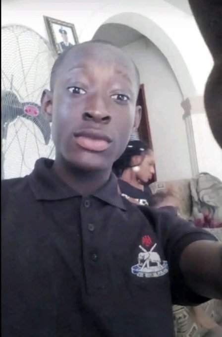 A young man in a black polo shirt with a crest takes a selfie in a room with an oscillating fan and framed picture in the background, with another person visible behind him.