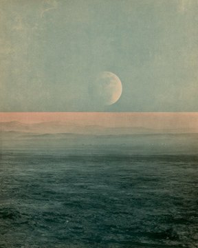 A serene, muted-toned photograph of a half moon rising over a calm ocean horizon with a soft pinkish band of light along the waterline.