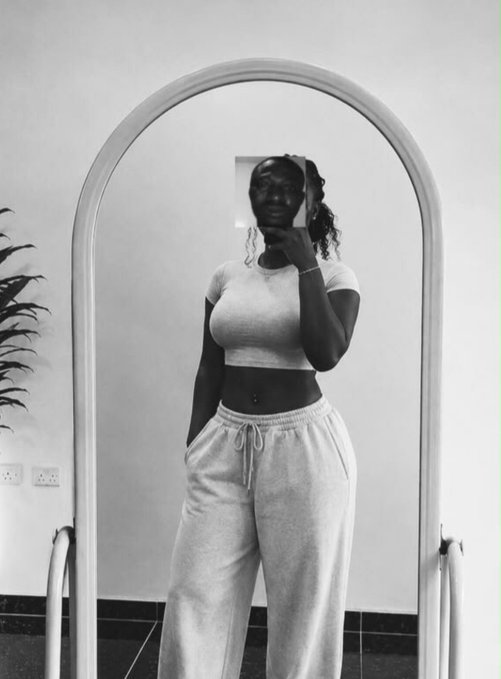 A black-and-white photo of a woman taking a mirror selfie while wearing a white short-sleeved crop top and gray sweatpants.