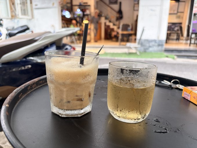 Two iced beverages—one a creamy coffee-like drink with a black straw, the other a pale, bubbly drink—sit on a small black tray at an outdoor café, with the partial text "LEAD" visible on a parked scooter in the background, setting a relaxed street-side scene fitting the celebratory "GM" message.