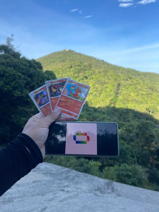 The image features a person holding up three Pokemon cards against a backdrop of a mountain range, with trees visible in the background. The person's hand is positioned over the top left corner of the photo, while the Pokemon cards are displayed on their right side. The text "Conquer the peaks with Renaiss! Bring the spirit of exploration from the digital world to the majestic wilderness." suggests that the person might be a fan of Pokemon and has used the cards as part of an adventure or exploration in nature.