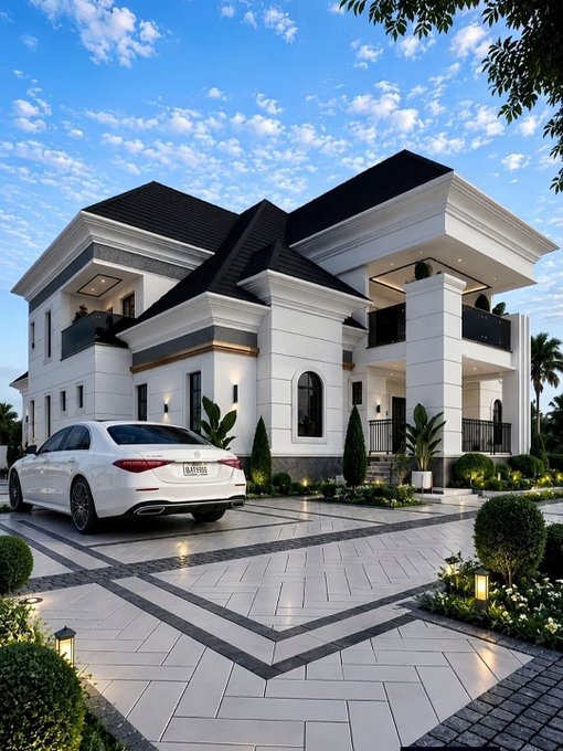 A modern white two-story house with black roofing is illuminated at dusk, featuring a geometric patterned driveway where a white luxury sedan is parked, surrounded by manicured landscaping.