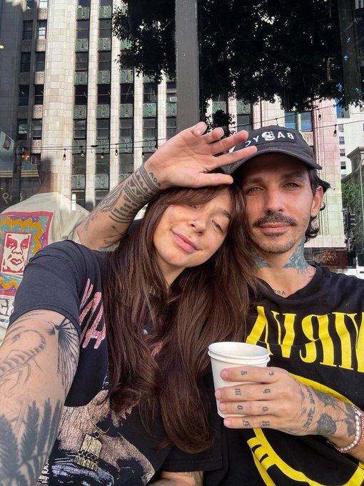The image features a man and woman posing together for a photo, with the woman holding a coffee cup while the man holds her hand. The background includes a building and trees, suggesting an urban setting. The caption of the image reads "Rocking that vintage @BoredApeYC hat today!". This implies that they are enjoying their time together in this location, possibly taking a break from their daily routines to spend quality time with each other.

The woman is wearing a black shirt and has tattoos on her arms, while the man is wearing a yellow shirt. They both have coffee cups in their hands, which adds to the casual and relaxed atmosphere of the photo. The presence of trees in the background further enhances the urban setting, making it feel more like an outdoor cafe or gathering spot.