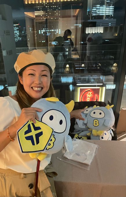 A woman in a white chef's hat beams while holding up a box of colorful character macarons, with more boxed macarons and plush toys stacked behind her at the BTCC event in GINZA SIX, matching the post's mention of the piled-up Nakamon-themed treats and original macarons.