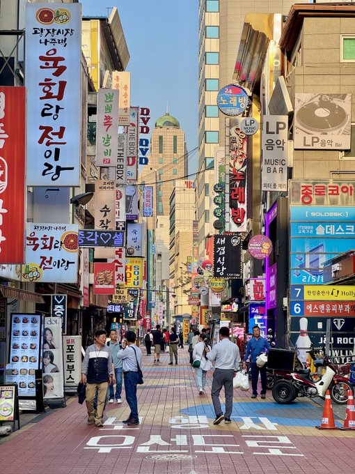 A bustling street scene in Korea featuring tall buildings covered in colorful signs with Korean text, including 'LP MUSIC BAR', and pedestrians walking along a brick-paved road.