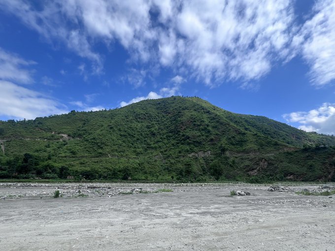 A wide landscape view shows a green forested mountain under a blue sky with scattered clouds, with a sandy, rocky riverbed in the foreground.