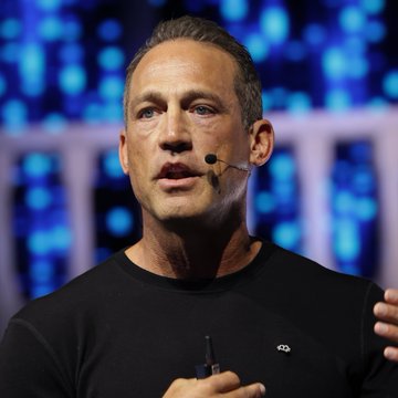 A man with short hair speaks on stage wearing a black t-shirt with a 'dg' logo and a headset microphone against a blue digital backdrop.