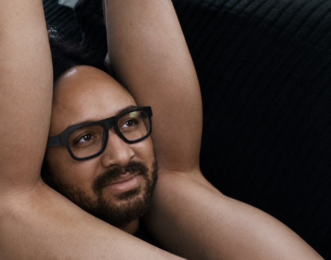 A portrait of a bearded man wearing black-framed glasses, posed with his arms raised behind his head against a dark background.