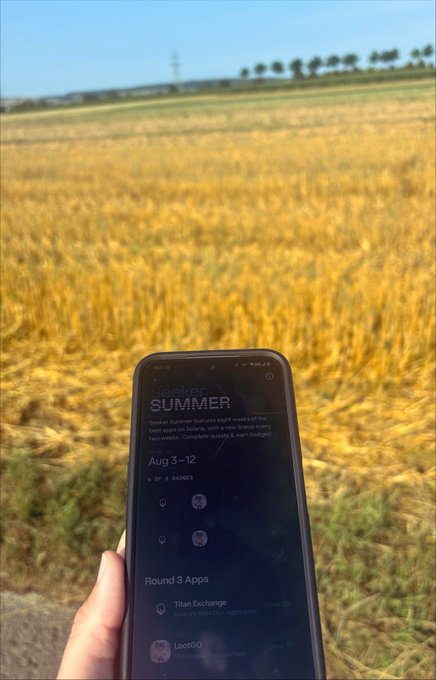 A hand holds a phone displaying the "Seeker SUMMER" app's "Aug 3-12" event page against a golden wheat field backdrop, with visible tasks including "Titan Exchange" and "Loot00" under "Round 3 Apps," perfectly capturing the post's vibe of earning Solana rewards during a scenic outdoor walk.