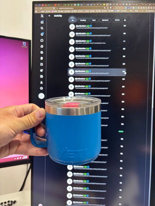 A hand holds a blue mug in front of a vertical monitor displaying a social media activity feed on the X platform.