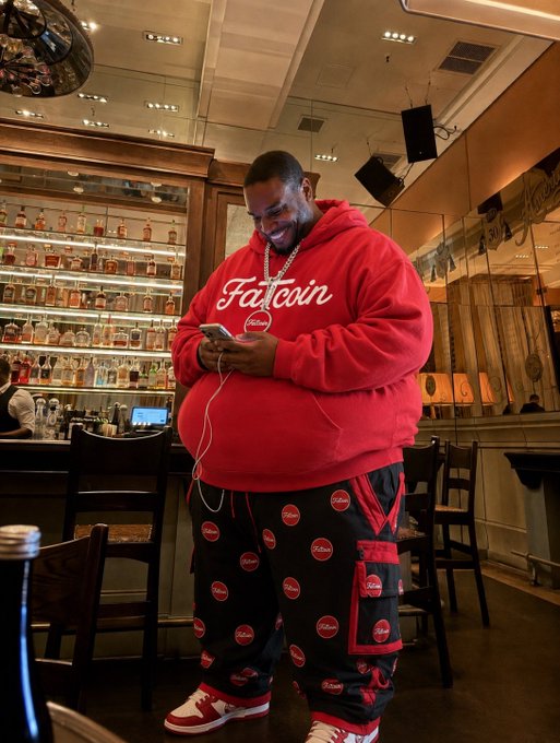 A man smiles while looking at his phone in what appears to be a bar or lounge setting, wearing a red hoodie prominently displaying "Fatcoin" branding and matching black cargo pants with repeating red Fatcoin logos, promoting the cryptocurrency meme coin referenced in the post.