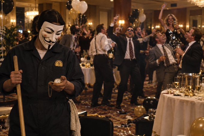 A person wearing a Guy Fawkes mask and dark coveralls holding a mop in the foreground, while a formal party with people in suits and dresses celebrating takes place in the background.