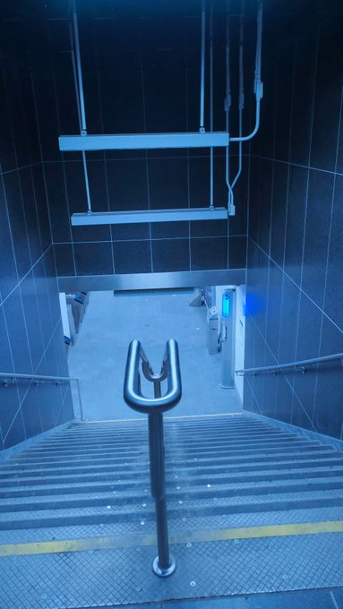 A dimly lit view looking down a staircase with metal grating steps and a central handrail, leading to a subway or industrial station entrance with a blue-lit interior at the bottom.