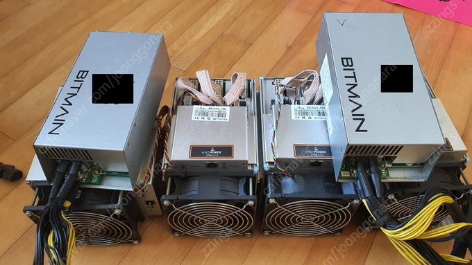 Two Bitmain ASIC miners, likely Antminer S9 units identifiable by the "BITMAIN" branding on their silver housings and control board labels, sit side-by-side on a wooden floor with cooling fans and power cables exposed—representing the early crypto mining operation described, where electricity costs drove a move to a cheaper factory location before expanding the setup into a node-rig.