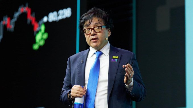 An Asian man in a blue suit, blue tie, and glasses speaks at a presentation, with a 'TOM LEE' name tag and a financial chart visible in the background.