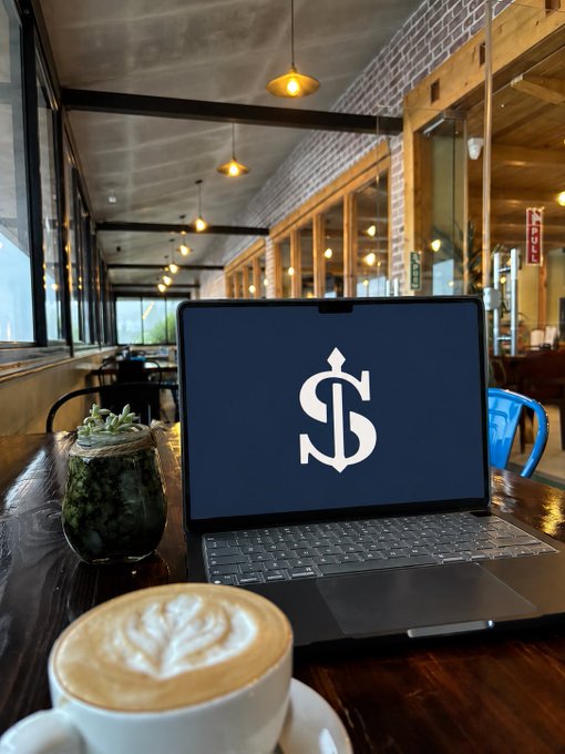 A laptop displaying a stylized dollar sign logo sits on a wooden table in a cafe, accompanied by a latte with leaf-shaped foam art and a small potted succulent.