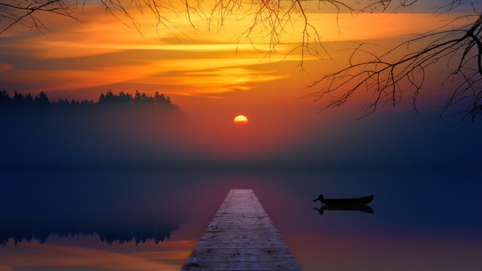 A vibrant orange and red sunrise over a foggy lake reflects onto the water, with a wooden dock and a small boat in the foreground.